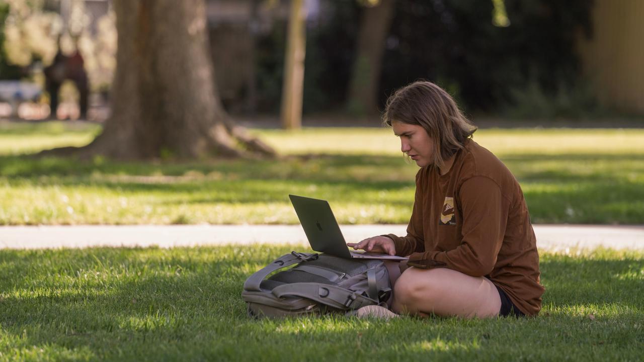 A student sitting on the grass with a laptop and backpack